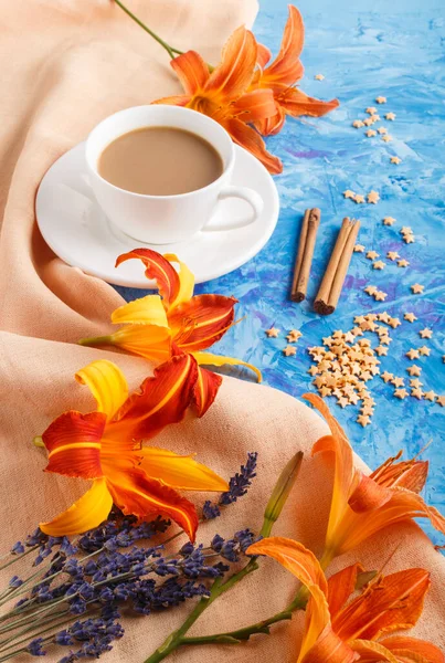 Orange day-lily and lavender flowers and a cup of coffee on a blue concrete background, with orange textile. Morninig, spring, fashion composition. side view.