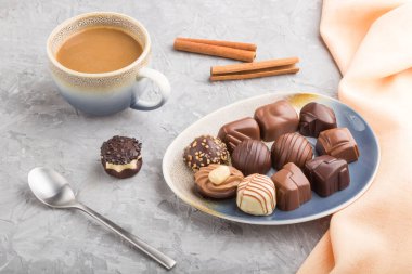 Farklı çikolata şekerleri ve gri beton arka planda bir fincan kahve ve turuncu tekstil. Yan görünüm, kapat