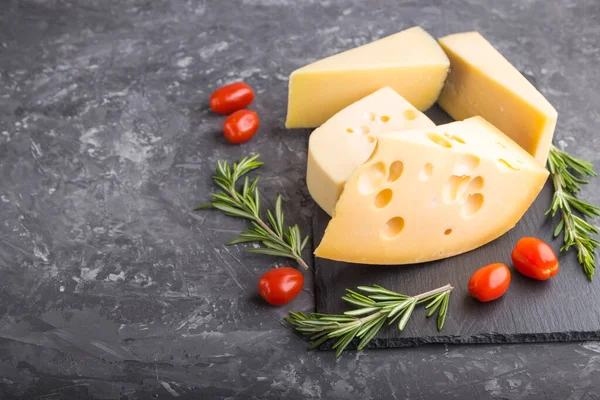 Various types of cheese with rosemary and tomatoes on black slate board on a black concrete background. Side view, close up, copy space, selective focus.