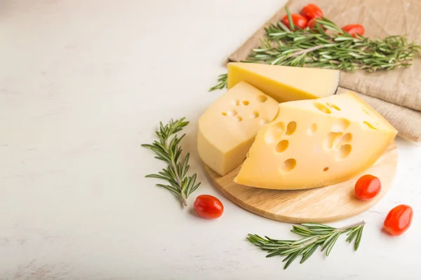 Various types of cheese with rosemary and tomatoes on wooden board on a white wooden background and linen textile. Side view, close up, copy space, selective focus.