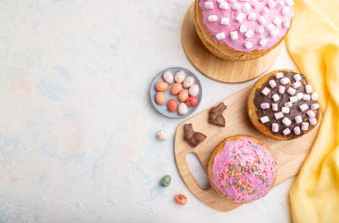 Beyaz beton arka plan ve sarı tekstil üzerinde çikolata yumurtaları ve tavşanlarla ev yapımı ve dekore edilmiş paskalya turtaları. Üst görünüm, düz görünüm, kopyalama alanı.