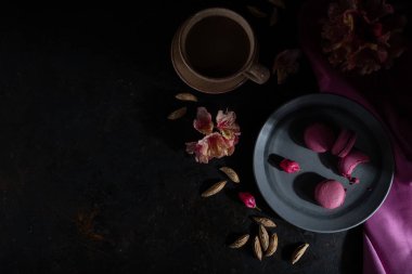 Mor badem kurabiyeleri ya da siyah beton arka planda bir fincan kahve ve pembe tekstil içeren badem kurabiyeleri. Sert ışık, sade anahtar. Üst görünüm, düz görünüm, kopyalama alanı.