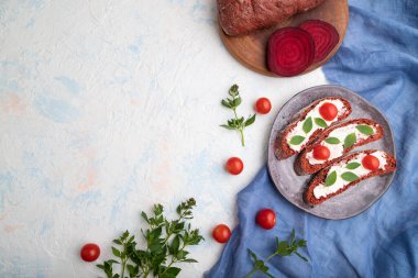 Krem peynirli kırmızı pancar ekmekli sandviçler ve beyaz beton arka planda domatesler ve mavi keten kumaş. Üst görünüm, düz görünüm, kopyalama alanı.