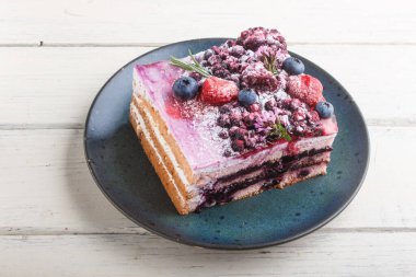 Süt kremalı böğürtlenli kek ve beyaz ahşap arka planda mavi seramik tabakta yabanmersini reçeli. Yan görünüm, kapat.