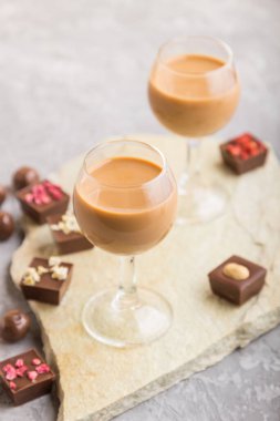 Bardakta çikolata likörü, gri beton arka planda çikolata şekeri ve taş levha. Yan görünüm, yakın görünüm, seçici odak.