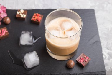 Bardakta buzlu çikolata likörü, gri beton arka planda çikolata şekeri ve siyah taş levha. Yan görünüm, yakın görünüm, seçici odak.