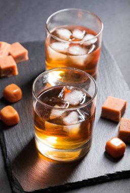 Bir bardak kehribar viskisi, buzlu ve karamel şekerli siyah arka planda siyah taş bir tahta üzerinde. Yan görünüm, yakın görünüm, seçici odak.