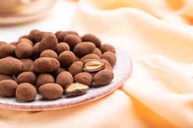 Seramik tabakta çikolatalı bardakta badem ve beyaz beton arka planda bir fincan kahve ve turuncu keten kumaş. Yan görünüm, yakın görünüm, seçici odak.