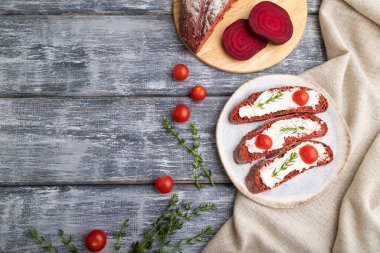 Krem peynirli kırmızı pancar ekmekli sandviç ve gri ahşap arka planda domates ve keten tekstil. Üst görünüm, düz görünüm, kopyalama alanı.