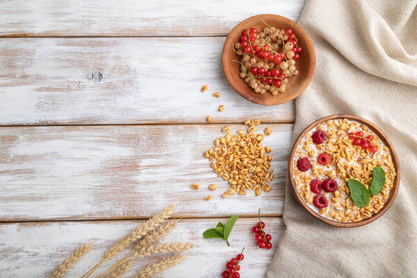 Wheat flakes porridge with milk, raspberry and currant in wooden bowl on white wooden background and linen textile. Top view, flat lay, copy space.