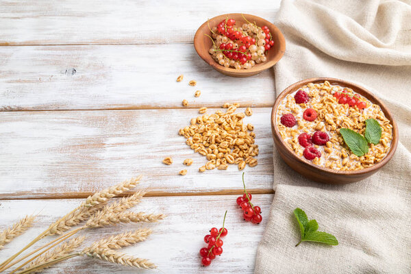 Wheat flakes porridge with milk, raspberry and currant in wooden bowl on white wooden background and linen textile. Side view, copy space.