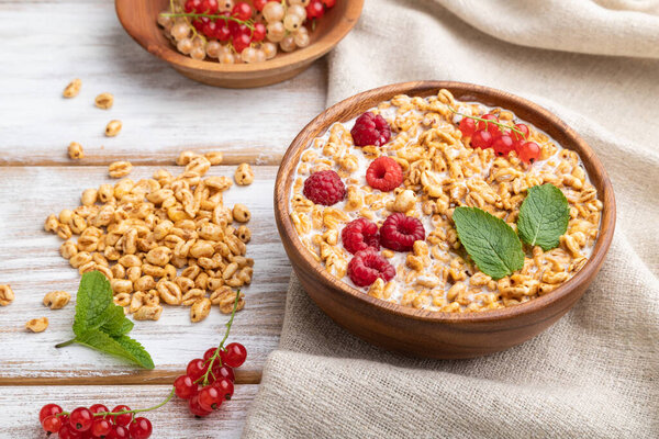 Wheat flakes porridge with milk, raspberry and currant in wooden bowl on white wooden background and linen textile. Side view, close up.