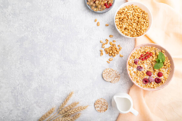 Wheat flakes porridge with milk, raspberry and currant in ceramic bowl on gray concrete background and orange linen textile. Top view, flat lay, copy space.