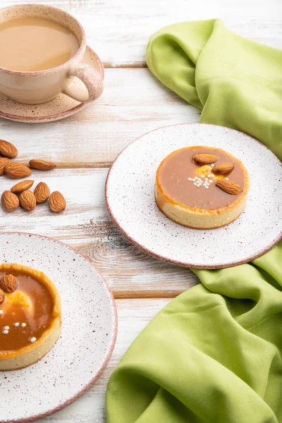 Sweet tartlets with almonds and caramel cream with cup of coffee on a white wooden background and green textile. side view, close up.