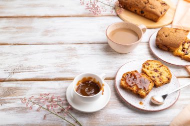 Kuru üzümlü, bademli, yumuşak karamel ve beyaz ahşap arka planda bir fincan kahve ve turuncu keten tekstil. Yan görünüm, boşluk kopyala.