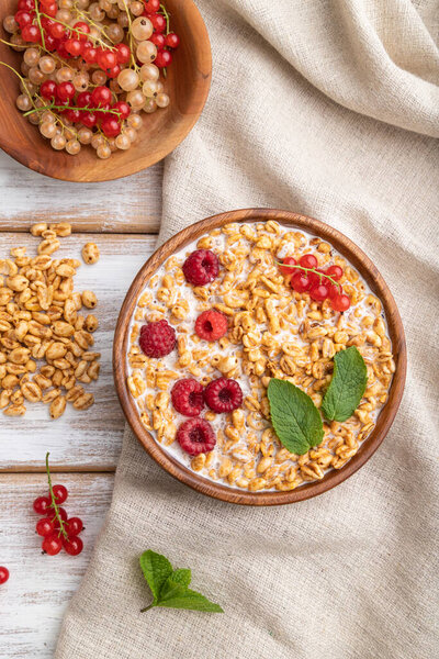 Wheat flakes porridge with milk, raspberry and currant in wooden bowl on white wooden background and linen textile. Top view, flat lay, close up.