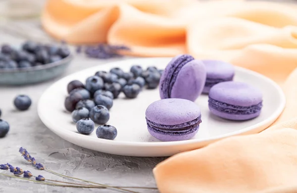 Purple macarons or macaroons cakes with blueberries on white ceramic plate on a gray concrete background and orange textile. Side view, close up, selective focus.