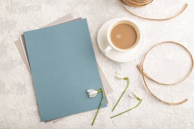 Bahar kardelen galanthus çiçekleri ve gri beton arka planda bir fincan kahveyle mavi kağıt maketi. Boşluk, kartvizit, üst manzara, düz yatak, yakın çekim, durgun hayat. bahar konsepti.