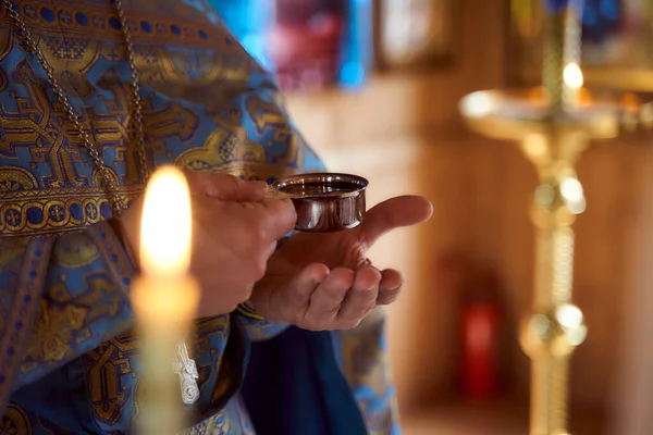 Las manos de los sacerdotes sostienen un tazón pequeño de vino en el ambiente de la iglesia. Velas en el fondo — Foto de Stock