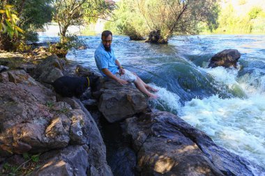 Genç bir adam fırtınalı bir dağ nehrinin kıyısında bir taşın üzerinde oturuyor.