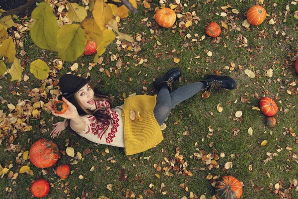 Güzel kız çimlerin üzerinde oturuyor. Üst Manzara. Elinde küçük dekoratif bir balkabağı tutuyor. Dilini uzatıyor..