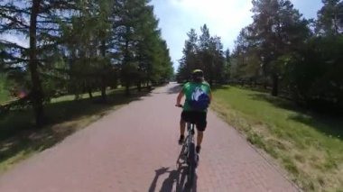 Parkta bisiklet sürmek. Bisikletçi ağaçlar arasında yol boyunca gidiyor. Kamera onu takip ediyor ve arkasından yakalıyor. Yaz. Güneşli hava.