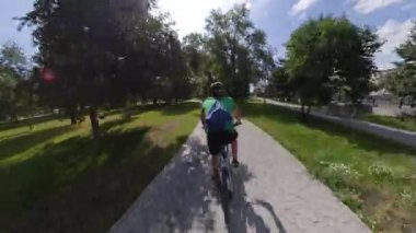 Parkta bisiklet sürmek. Bisikletçi ağaçlar arasında yol boyunca gidiyor. Kamera onu takip ediyor ve arkasından yakalıyor. Yaz. Güneşli hava.