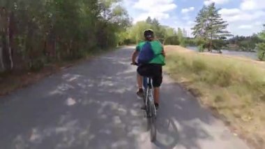 Parkta bisiklet sürmek. Bisikletçi ağaçlar arasındaki patika boyunca hızla gider. Kamera onu takip ediyor ve arkasından yakalıyor. Yaz. Güneşli hava.