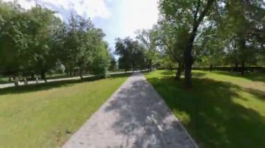 Yazın küçük bir kasaba parkı. Kamera parkta patika boyunca hızlı hareket eder. Ağaçlar, yeşil çimenler ve güneşli bir gökyüzü görülebilir.