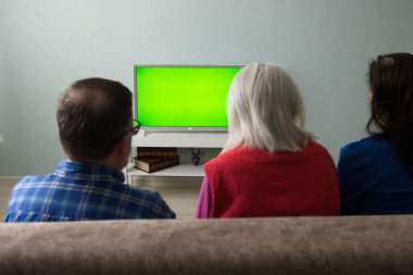 Aile televizyon izliyor. Yeşil ekran. Üç orta yaşlı ve yaşlı insan kanepede oturuyor. Önlerinde yeşil ekran bir televizyon var.. 