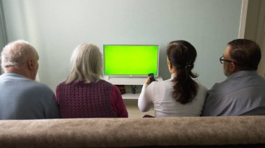 Aile televizyon izliyor. Yeşil ekran. İki çift, bir yaşlı ve orta yaşlı çift, evdeki kanepede oturuyorlar. Önlerinde yeşil ekran bir televizyon var. Kadın televizyonun uzaktan kumandasına basıyor..