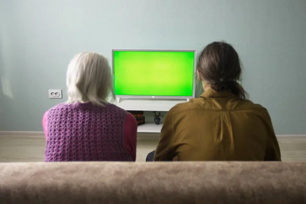 Aile televizyon izliyor. Yeşil ekran. Büyükanne ve yetişkin torun kanepede oturuyorlar. Önlerinde yeşil ekran bir televizyon var.. 