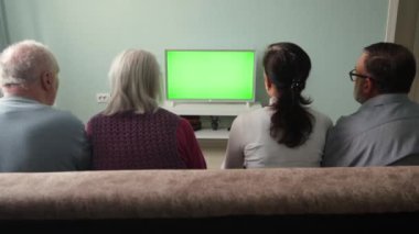 Aile televizyon izliyor. Yeşil ekran. İki çift, bir yaşlı ve orta yaşlı çift, evdeki kanepede oturuyorlar. Önlerinde yeşil ekran bir televizyon var. Kadın televizyonun uzaktan kumandasına basıyor..