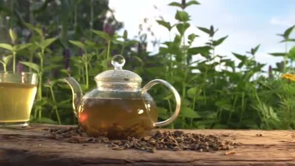 自然の中でお茶 スローモーション2倍 花の咲くミントを背景に緑茶との静物 テーブルの上でお茶 乾燥茶 シャモアを醸造しています 男の手はお茶のマグカップを取ります — ストック動画