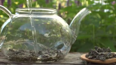 Doğada çay. Ağır çekim 4x. Kaynar su yeşil çay yapraklarıyla bir çaydanlığa dökülür. Arka planda nane yetişir. Kameranın yatay hareketi.