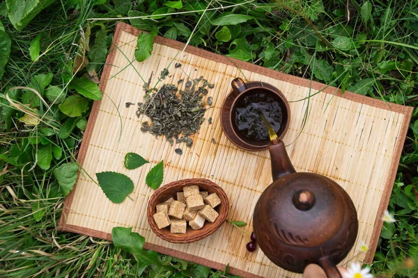 Tea in nature. Flat lay. On a bamboo napkin, dry and brewed green tea, mint leaves, a couple of cherries and cane sugar cubes.A man\'s hand pours tea from a teapot into a cup.