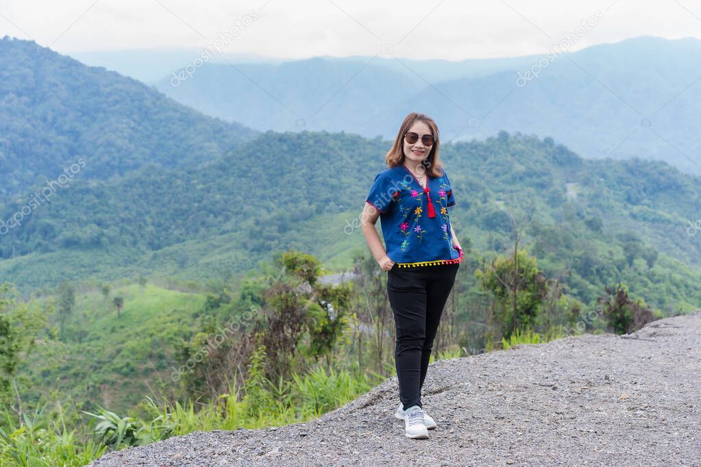 Adult traveling woman wearing sunglasses standing and smiling on the top of the mountain cliff with relaxing mood and watching beautiful view of woods and clouds foggy on vacation. Nature background