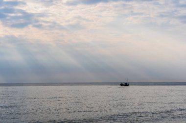 Güneşin altın ışıklarıyla aydınlatılan parlak deniz ve bulutların manzarası. Bulutların arkasından küçük bir balıkçı teknesinin silüetiyle çıkıyor. Sabah balığa çıkmak üzereler..