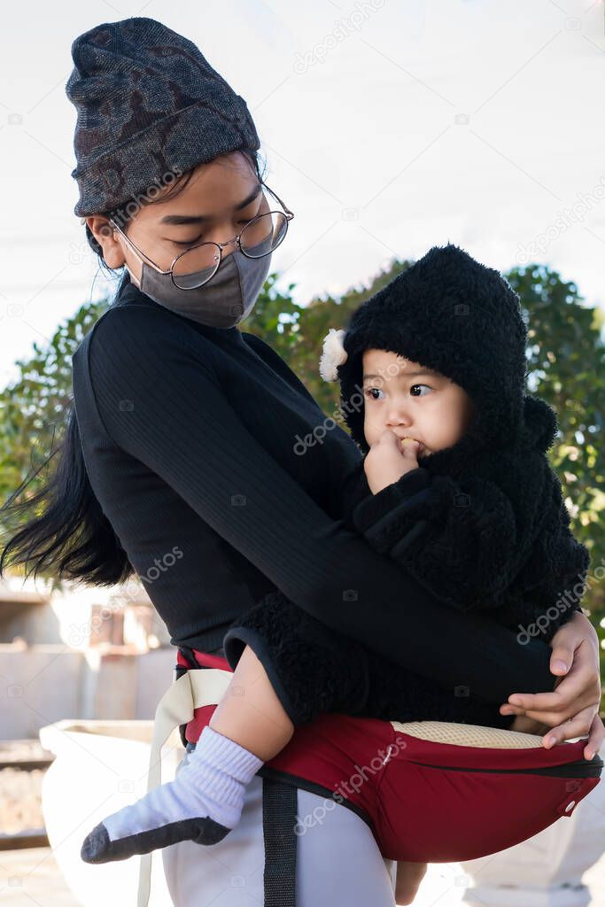 Portrait of Asian mother wears protective mask and holds her adorable young daughter. Child girl eating sweets with dirty mouth. Healthcare and social distancing for the Covid-19 epidemic concep