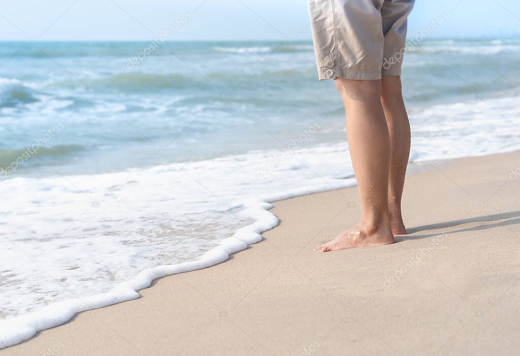 Young woman's bare feet with a wave coming and enjoying traveling to exotic nature. Beautiful healthy female legs with droplets standing barefoot alone on sandy sea beach in summer. Lifestyle concept