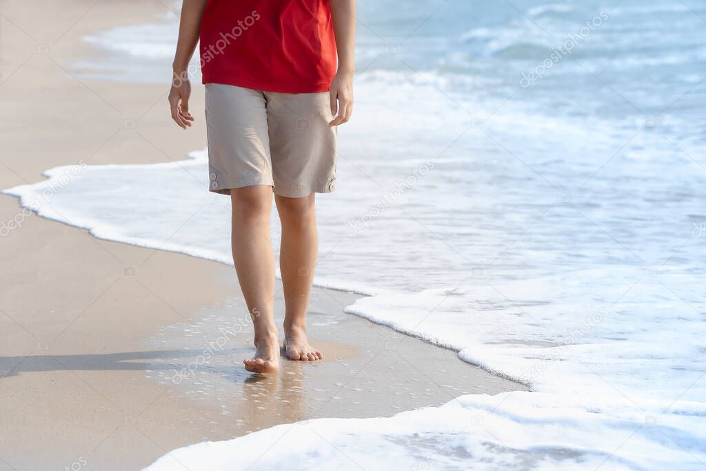 Young woman's bare feet with a wave coming and enjoying to travel on tropical beach in sunlight. Healthy female tan legs with droplets walking barefoot alone on sandy sea beach in summer. Copy space.