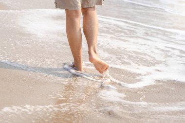 Genç bir kadının çıplak ayaklarıyla ayaklarına gelen dalgalarla. Damlacıkları olan güzel, sağlıklı kadın bacakları kumlu sahilde yalınayak yürüyor ve yazın tropik plajlarda gezmenin tadını çıkarıyor.