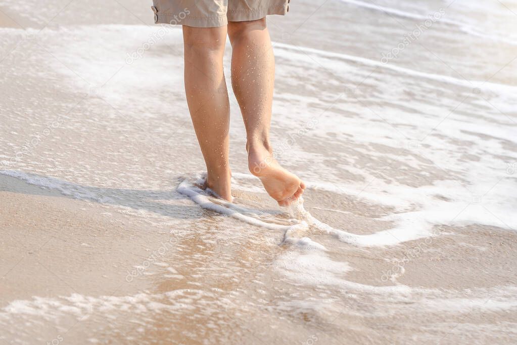 Young woman's bare feet with wave motion coming to the foot. Beautiful healthy female legs with droplets walking barefoot alone on sandy sea beach and enjoying to travel tropical beach in summer
