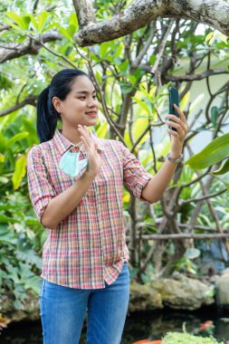 Güzel Asyalı genç bir kadın akıllı telefonu elinde tutuyor. Arkadaşlarını, sevgililerini ya da ailesini aramak için cep telefonu uygulaması kullanıyor. Bahçede elinde tıbbi maske tutuyor..
