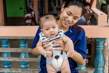 Genç bir kadının kolundaki ilgiyle kameraya bakan sevimli çocuk. Asyalı bir kadın kendi bebeğini tutarken, küçük Asyalı bir bebeğin anne ve babasına bakması. Mutlu bebeğe odaklan. 