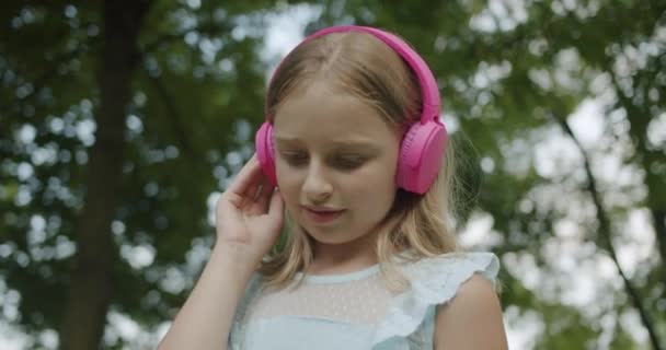 Niño pequeño escucha música en los auriculares rosados modernos en el parque y mira hacia abajo — Vídeos de Stock