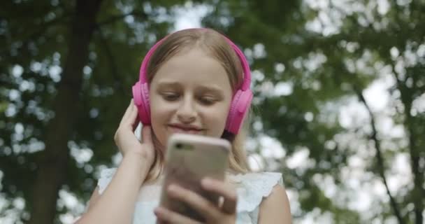 Feliz adolescente escucha música en modernos auriculares rosados con teléfono móvil — Vídeo de stock