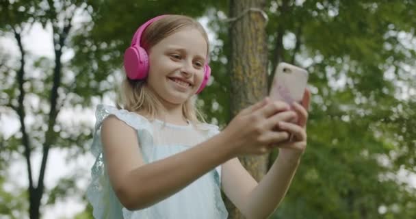 Happy Girl comunica videollamada en línea con auriculares rosados modernos en el parque — Vídeos de Stock