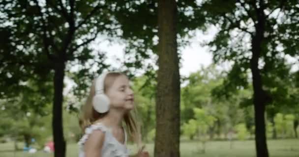 Das fröhliche Kind, das zu Musik tanzt, springt mit Kopfhörern im Park auf — Stockvideo