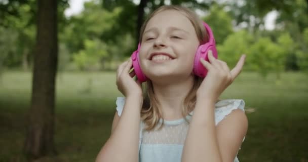 Adolescente feliz escucha música en los auriculares rosados modernos en el parque y mira hacia arriba — Vídeos de Stock